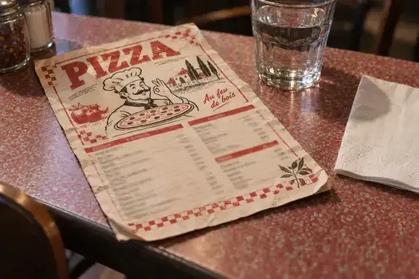 Un flyer de pizzeria des ann&eacute;es 90 pos&eacute; sur une table en formica, &agrave; c&ocirc;t&eacute; d'un verre d'eau
